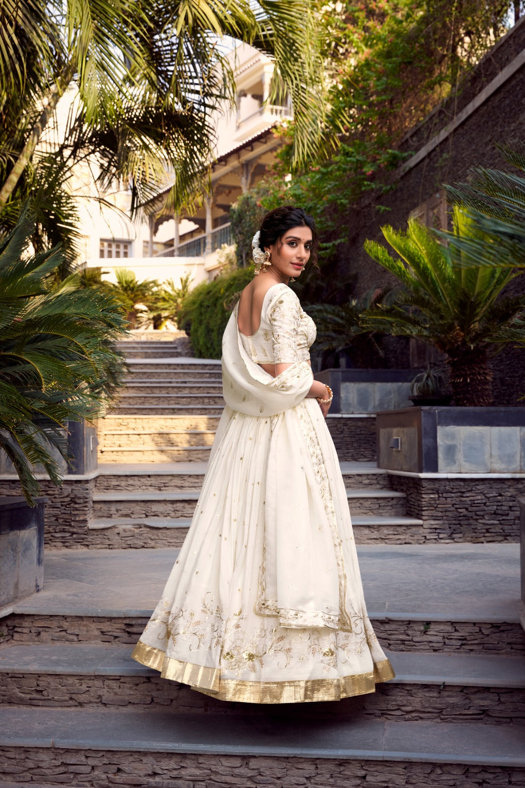 Woman in a white traditional outfit Lehenga Choli standing on stone steps with greenery in the background